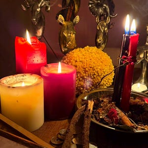 Pode incluir: Uma cole&ccedil;&atilde;o de velas acesas em vermelho, rosa e branco, ao lado de uma tigela de metal cheia de bot&acirc;nicos secos, pequenas moedas e dois gravetos. Atr&aacute;s, tr&ecirc;s esculturas met&aacute;licas de cabe&ccedil;as de animais, com o texto KART & KITAP.