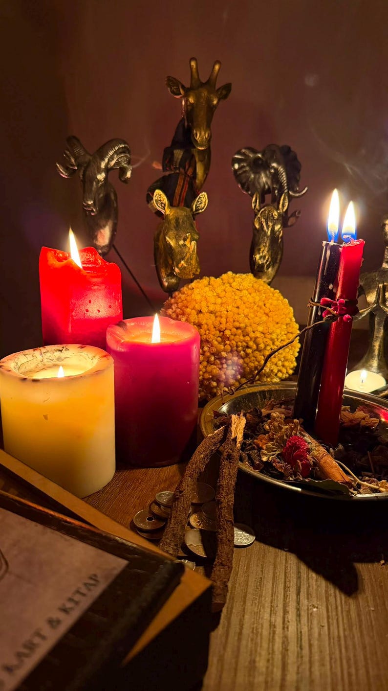 Pode incluir: Uma cole&ccedil;&atilde;o de velas acesas em vermelho, rosa e branco, ao lado de uma tigela de metal cheia de bot&acirc;nicos secos, pequenas moedas e dois gravetos. Atr&aacute;s, tr&ecirc;s esculturas met&aacute;licas de cabe&ccedil;as de animais, com o texto KART & KITAP.