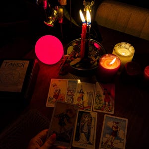 Pode incluir: Uma cole&ccedil;&atilde;o de cartas de tar&ocirc; espalhadas sobre uma superf&iacute;cie escura, incluindo uma carta de Joker e uma carta com a etiqueta Azize. Uma vela vermelha, uma branca, um orbe rosa brilhante e uma ta&ccedil;a de metal com velas est&atilde;o dispostos.