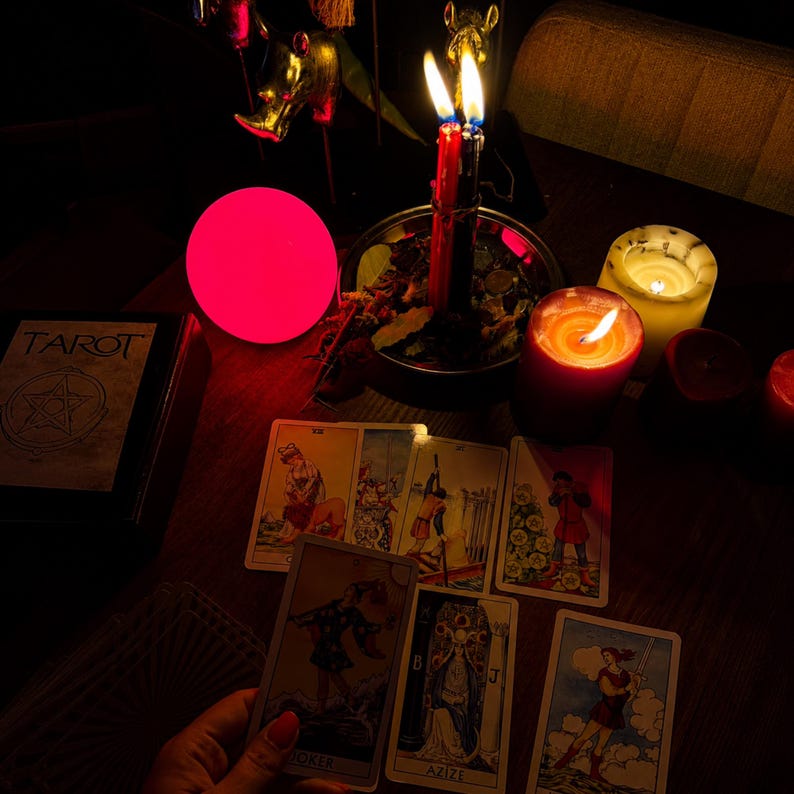 Pode incluir: Uma cole&ccedil;&atilde;o de cartas de tar&ocirc; espalhadas sobre uma superf&iacute;cie escura, incluindo uma carta de Joker e uma carta com a etiqueta Azize. Uma vela vermelha, uma branca, um orbe rosa brilhante e uma ta&ccedil;a de metal com velas est&atilde;o dispostos.