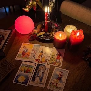 Pode incluir: Uma disposi&ccedil;&atilde;o de sete cartas de tarot sobre uma superf&iacute;cie de madeira, incluindo cartas com as etiquetas G&Uuml;&Ccedil;, İ&Ccedil; KRAL, AY, AZİZE e KILI&Ccedil; PRENSİ. A cena &eacute; iluminada por uma esfera rosa brilhante, duas velas acesas numa ta&ccedil;a de metal e duas velas de pilar.