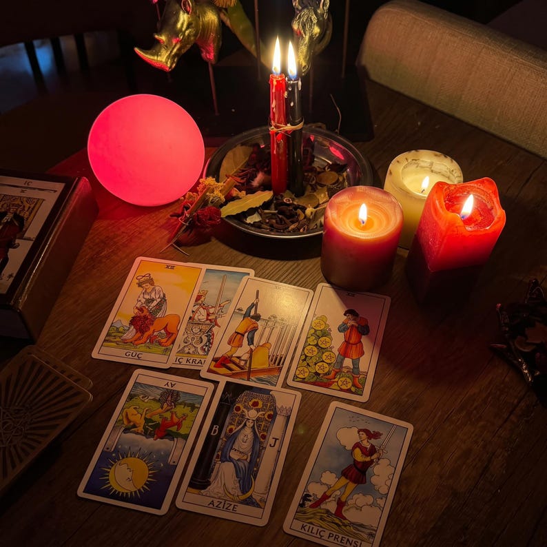 Pode incluir: Uma disposi&ccedil;&atilde;o de sete cartas de tarot sobre uma superf&iacute;cie de madeira, incluindo cartas com as etiquetas G&Uuml;&Ccedil;, İ&Ccedil; KRAL, AY, AZİZE e KILI&Ccedil; PRENSİ. A cena &eacute; iluminada por uma esfera rosa brilhante, duas velas acesas numa ta&ccedil;a de metal e duas velas de pilar.
