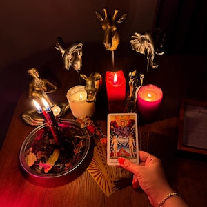 Pode incluir: Uma m&atilde;o segura uma carta de tarot dos Amantes com duas figuras sob um anjo. A cena inclui figuras de animais douradas, uma vela pilar vermelha, uma vela rosa, uma branca e uma tigela de metal cheia de folhas secas e duas velas acesas.