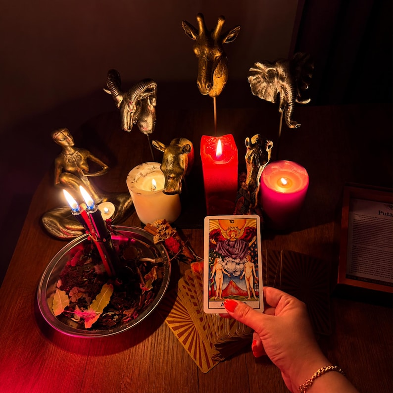Pode incluir: Uma m&atilde;o segura uma carta de tarot dos Amantes com duas figuras sob um anjo. A cena inclui figuras de animais douradas, uma vela pilar vermelha, uma vela rosa, uma branca e uma tigela de metal cheia de folhas secas e duas velas acesas.