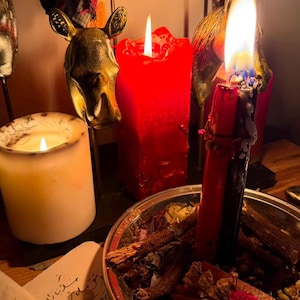 Pode incluir: Uma vela vermelha e preta acesa repousa numa ta&ccedil;a de metal cheia de ervas e flores secas. Perto, uma vela branca arde, rodeada por figuras met&aacute;licas de animais, incluindo um elefante e um rinoceronte. Um papel com escrita &aacute;rabe encontra-se em primeiro plano.