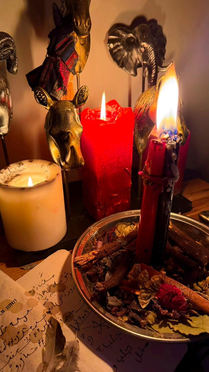 Pode incluir: Uma vela vermelha e preta acesa repousa numa ta&ccedil;a de metal cheia de ervas e flores secas. Perto, uma vela branca arde, rodeada por figuras met&aacute;licas de animais, incluindo um elefante e um rinoceronte. Um papel com escrita &aacute;rabe encontra-se em primeiro plano.