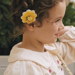 Könnte beinhalten: Eine gelbe Blumen-Haarspange mit weißen, gewellten Rändern und einer strukturierten Mitte mit kleinen gelben und grünen Details. Das Accessoire ist an der Seite einer geflochtenen Frisur befestigt und zeigt ein Design mit geschichteten Blütenblättern.