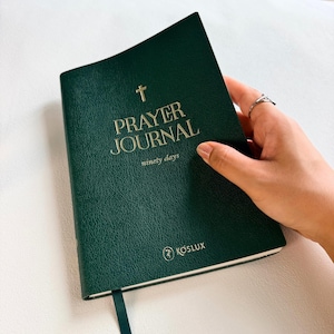 Könnte beinhalten: Ein dunkelgrünes Gebetstagebuch mit strukturiertem Einband, verziert mit einem goldenen Kreuzsymbol und dem in Gold gedruckten Text PRAYER JOURNAL ninety days. Unten auf dem Einband befinden sich ein kleines kreisförmiges Logo und der Markenname KOSLUX.