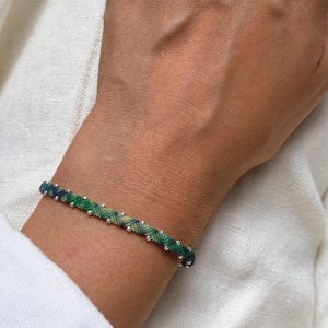 Può includere: Un braccialetto intrecciato caratterizzato da un mix di fili verdi e blu scuro intrecciati insieme, accentuato da piccole perline metalliche color argento distanziate uniformemente lungo l'intera lunghezza del cordino, creando un accessorio dettagliato.