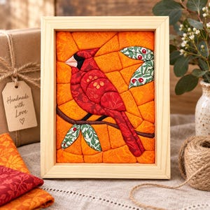 May include: A framed fabric mosaic art piece featuring a red cardinal bird perched on a brown branch against an orange background with leaf patterns. The bird is constructed from various red patterned fabric segments, with green leaf accents.