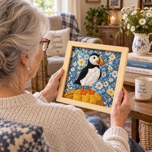 Könnte beinhalten: Ein quadratischer Holzrahmen mit einem gesteppten Stoffkunstwerk. Das Quilt zeigt einen Papageientaucher mit schwarz-weißem Körper, orangefarbenem Schnabel und orangefarbenen Füßen, der auf einem gelben Felsen vor blauem Blumenhintergrund steht.