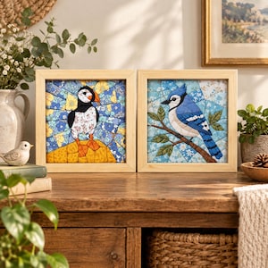 Op de afbeelding: Twee vierkante ingelijste stoffen kunstwerken met gewatteerde vogelontwerpen. Het linkerframe toont een papegaaiduiker op een gele ondergrond met blauwe bloemaccenten. Het rechterframe toont een blauwe gaai op een bruine tak tegen een blauwe bloemenachtergrond.