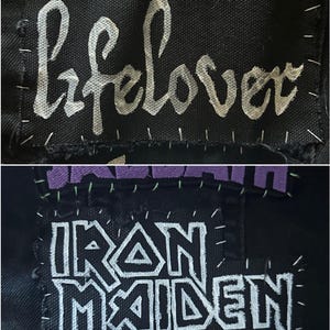 May include: A black fabric patch featuring the white text lifelover in a cursive font, hand-stitched with silver thread. Below it, a black fabric patch displays the white text Iron Maiden in a block font, also hand-stitched with silver thread.
