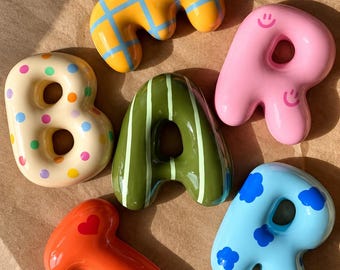May include: Five ceramic letters forming the word BAR, plus an additional letter T. The letters feature various designs: a yellow grid, pink with smiley faces, cream with colorful polka dots, olive green with white stripes, orange with a red heart, and blue with clouds.