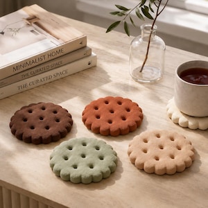 May include: A set of four round, scalloped-edge coasters shaped like biscuits in dark brown, terracotta, sage green, and beige. Each coaster features a textured surface with small indentations, mimicking the appearance of a classic round biscuit.
