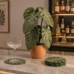 Puede incluir: Un juego de posavasos verdes con forma de hoja de monstera y un soporte a juego con forma de planta. Los posavasos son planos con detalles de nervaduras blancas, mientras que el soporte presenta una maceta naranja con patrón en espiral.