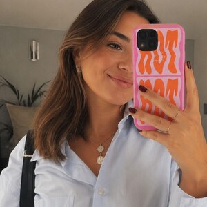 May include: A light blue button-down shirt with a chest pocket and a pink smartphone case featuring orange wavy text that reads HOT MOM. The phone case has a rectangular design with a black camera cutout and a glossy finish on the protective cover.