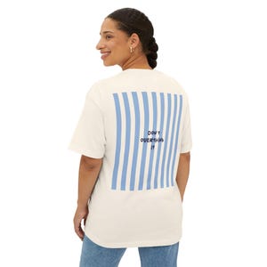 Puede incluir: Una camiseta de manga corta color blanco roto con un gran gráfico rectangular en la espalda con rayas verticales azul claro. Centrado entre las rayas, el texto azul oscuro "Don't overthink it" aparece en una fuente manuscrita informal.