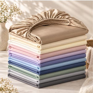 May include: A stack of folded bed sheets in various colors including champagne, cream, pale pink, lavender, light blue, navy, sage green, light gray, and dark charcoal. The top sheet is a fitted sheet with an elasticized edge in champagne.