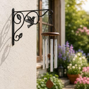 May include: A black metal wall bracket featuring a hummingbird and leaf design, supporting a wind chime with a circular wooden top, six silver-toned metal tubes of varying lengths, a central wooden striker, and a wooden teardrop-shaped wind catcher.