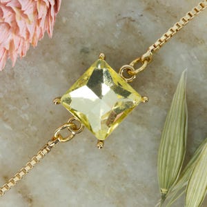 May include: A gold-toned box chain bracelet featuring a central square-cut, pale yellow gemstone held by four prongs. The delicate metal chain connects to the stone setting on opposite sides, creating a symmetrical and refined jewellery piece.