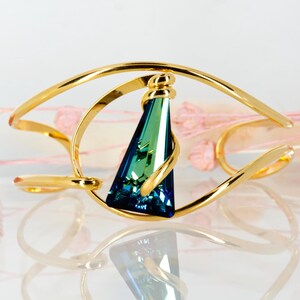 May include: A gold-toned cuff bracelet featuring a central, elongated triangular crystal that transitions in colour from deep blue at the base to a vibrant emerald green at the top, held within a curved, open-frame metal design.