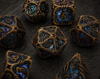 May include: A set of seven polyhedral dice featuring an antique gold-toned metal frame with intricate, swirling raised patterns. The recessed areas of each die are filled with a shimmering, iridescent blue and purple material with visible numbers.