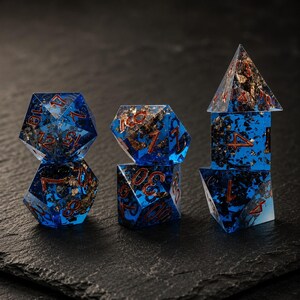 May include: A set of seven polyhedral dice made of translucent blue resin with embedded gold-coloured metallic flakes. Each die features engraved numbers filled with a deep red pigment, arranged in three stacks on a dark, textured surface.