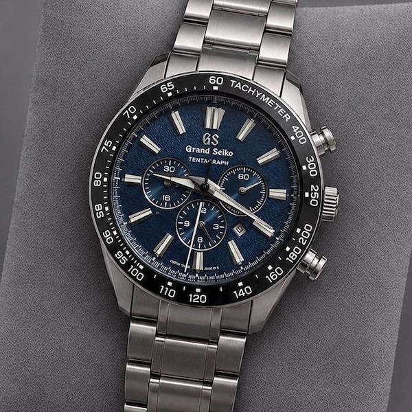 May include: A Grand Seiko Tentagraph chronograph watch with a textured blue dial, silver-tone hands and indices, and a black ceramic tachymeter bezel. The timepiece features a stainless steel link bracelet and a date display window.