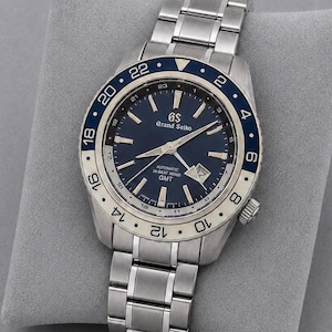Op de afbeelding: Een roestvrijstalen Grand Seiko automatisch horloge met een donkerblauwe wijzerplaat, zilverkleurige wijzers en indexen, en een datumvenster op de vier-uur-positie. Het horloge heeft een tweekleurige blauwe en zilveren GMT-ring met 24-uursmarkeringen.