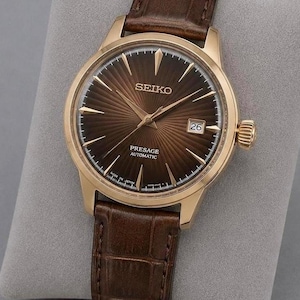 Op de afbeelding: Een Seiko Presage automatisch horloge met een roségoudkleurige kast en een donkerbruine wijzerplaat met zonnestraalpatroon. Het horloge heeft goudkleurige wijzers en uurmarkeringen, een datumvenster op de drie-uurpositie en een bruine leren band.