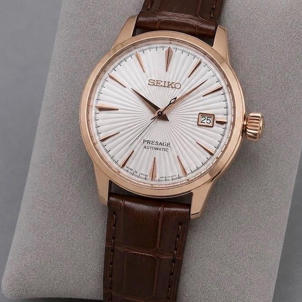 May include: A Seiko Presage Automatic watch with a rose gold-tone case, a white sunray-patterned dial, and rose gold-tone indices and hands. The watch features a date window at the three o'clock position and a brown leather strap with a crocodile texture.