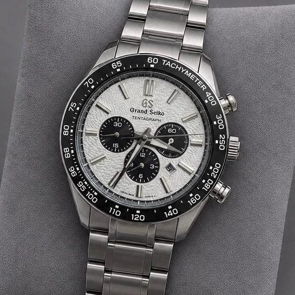 May include: A silver-toned Grand Seiko Tentagraph chronograph watch featuring a textured white dial, three black sub-dials, and a black ceramic tachymeter bezel. The timepiece includes a stainless steel link bracelet and silver hour markers.