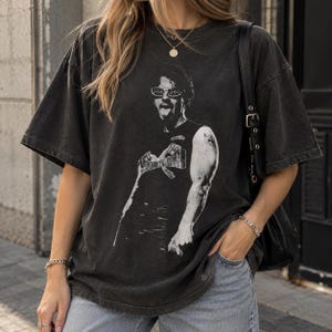 May include: A charcoal grey short-sleeve t-shirt featuring a black and white graphic print of a person wearing sunglasses and a tank top with the text JOE JOE. The shirt has a crew neckline, dropped shoulders, and a loose, relaxed fit.