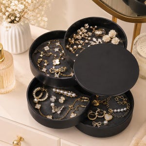 May include: A matte black rotating jewelry organizer with four circular tiers. The interior of each tray is lined with dark gray fabric, holding various gold-toned necklaces, bracelets, earrings, and pins featuring floral, butterfly, and pearl accents.
