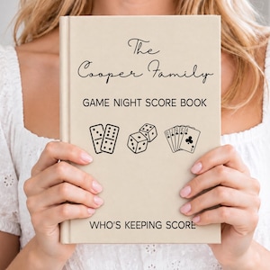 May include: A beige hardcover book titled The Cooper Family Game Night Score Book with black text and illustrations of dominoes, dice, and playing cards on the front cover, featuring the phrase Who's Keeping Score at the bottom of the cover.