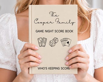 Può includere: Un libro con copertina rigida beige intitolato The Cooper Family Game Night Score Book con testo nero e illustrazioni di domino, dadi e carte da gioco sulla copertina, che include la frase Who's Keeping Score nella parte inferiore.