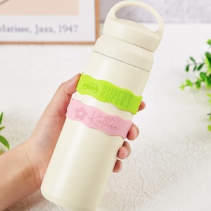 Pu&ograve; includere: Una borraccia termica color crema con un manico sul tappo. Due fasce in silicone ondulate avvolgono la bottiglia: una fascia verde lime con l'icona di un'auto e il nome RUPERT, e una fascia rosa chiaro con un fiore e il nome Kathie.