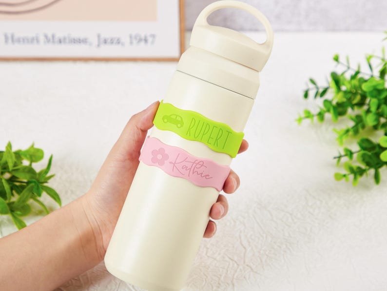 Pu&ograve; includere: Una borraccia termica color crema con un manico sul tappo. Due fasce in silicone ondulate avvolgono la bottiglia: una fascia verde lime con l'icona di un'auto e il nome RUPERT, e una fascia rosa chiaro con un fiore e il nome Kathie.