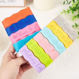 Pu&ograve; includere: Una collezione di dieci braccialetti in silicone ondulati e colorati con nomi personalizzati e varie icone tra cui un pallone da basket, una stella, un razzo, un trattore, una faccina sorridente e un cuore. I braccialetti sono arancioni, blu, rosa, azzurri, gialli e viola.