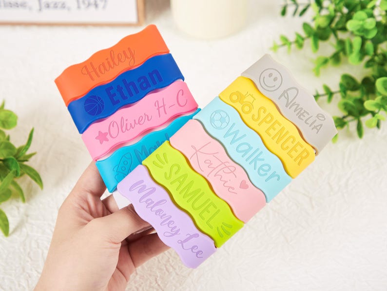 Pu&ograve; includere: Una collezione di dieci braccialetti in silicone ondulati e colorati con nomi personalizzati e varie icone tra cui un pallone da basket, una stella, un razzo, un trattore, una faccina sorridente e un cuore. I braccialetti sono arancioni, blu, rosa, azzurri, gialli e viola.