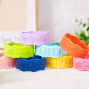 Pu&ograve; includere: Una collezione di braccialetti in silicone dai bordi ondulati, personalizzati con nomi e icone. I braccialetti si presentano in tonalit&agrave; lavanda, verde lime, azzurro, arancione, rosa, blu scuro e giallo, ognuno inciso con un testo unico.