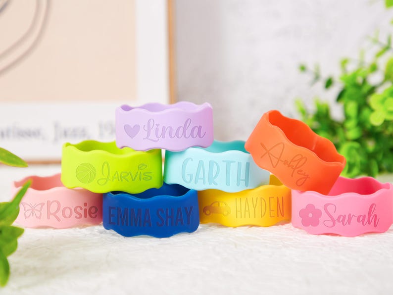 Pu&ograve; includere: Una collezione di braccialetti in silicone dai bordi ondulati, personalizzati con nomi e icone. I braccialetti si presentano in tonalit&agrave; lavanda, verde lime, azzurro, arancione, rosa, blu scuro e giallo, ognuno inciso con un testo unico.
