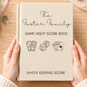 以下が含まれることがあります： ベージュのハードカバーのスコアブック。黒い文字で「The Foster Family」「GAME NIGHT SCORE BOOK」「WHO'S KEEPING SCORE」と書かれています。表紙には、2つのドミノ、2つのサイコロ、トランプの黒い線画が描かれています。