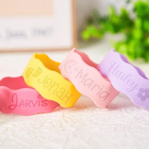 Pu&ograve; includere: Quattro braccialetti ondulati in silicone nei colori rosa, giallo, rosa chiaro e lavanda. Il braccialetto rosa riporta il nome Jarvis. Quello giallo mostra impronte di piedi di neonato e il nome Jonah. Il rosa chiaro presenta una luna, stelle e il nome Maria. Il braccialetto lavanda mostra il nome Hailey e un'icona a forma di fiore.
