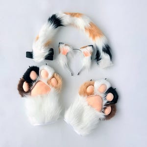 May include: A calico-patterned faux fur set consisting of a long, curved tail with a black elastic attachment, a headband featuring two pointed ears with white and orange fur, and two paw-shaped gloves with white fur and pink pads.