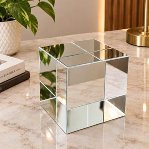 Pode incluir: Uma caixa decorativa quadrada construída a partir de múltiplos painéis de espelho reflexivos. O objeto apresenta uma disposição em grade de superfícies de vidro no topo e nas laterais, criando uma aparência multifacetada que reflete o ambiente.