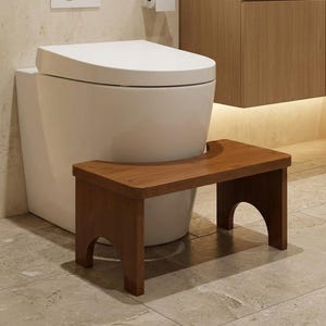 May include: A rectangular wooden step stool with a dark brown finish, featuring a curved cutout on the top surface and arched openings at the base of each side, positioned directly in front of a white toilet in a bathroom setting.