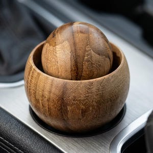 May include: A smooth, round wooden sphere resting inside a matching circular wooden bowl. Both items feature a natural brown wood grain pattern with visible textures and dark brown streaks, showcasing a polished finish and rounded edges.