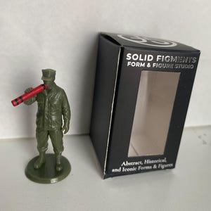 May include: An olive green plastic soldier figurine standing on a circular base, holding a red crayon with black stripes to its mouth. Beside it is a black rectangular box labeled SOLID FIGMENTS FORM & FIGURE STUDIO with a clear window.